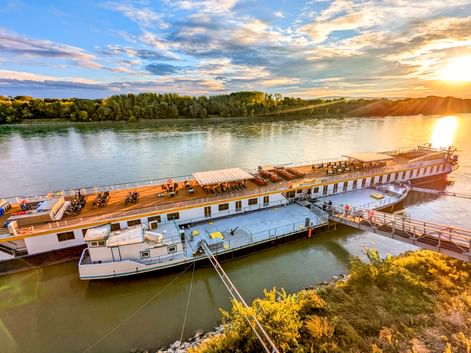 MS Prinzessin Katharina Flusskreuzfahrtschiff an der Donau bei Sonnenuntergang. Das weiße Schiff hat ein Holzdeck mit Fahrrädern und Liegestühlen.