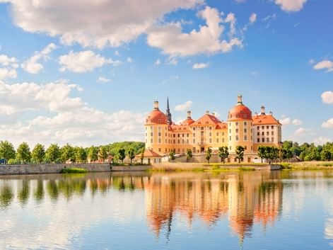 Schloss Moritzburg mit gelber Fassade und roten Kuppeln spiegelt sich im umgebenden See unter blauem Himmel mit weißen Wolken.