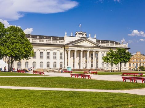 Das neoklassizistische Fridericianum in Kassel mit Säulenfassade, gesehen über eine grüne Rasenfläche mit roten Bänken unter blauem Himmel.