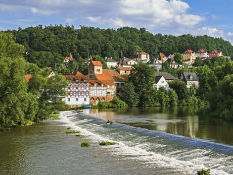 Blick auf Melsungen an der Fulda mit einem Wehr im Vordergrund. Fachwerkhäuser und rote Dächer schmiegen sich zwischen grüne Bäume unter blauem Himmel.