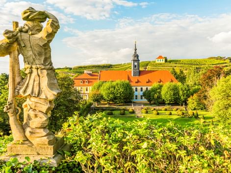 Steinstatue im Vordergrund mit Blick auf Gärten und historische Gebäude mit orangen Dächern in Diesbar-Seußlitz, dahinter weinbergbedeckte Hügel.