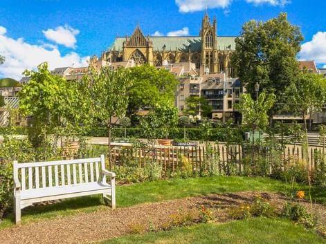 Weiße Bank im Garten mit Metzer Kathedrale hinter Wohngebäuden. Grüne Bäume und Rasen im Vordergrund unter blauem Himmel.