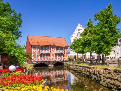 Rotes Fachwerkhaus mit orangefarbenem Dach über einem Kanal in Wismar. Bunte Blumen im Vordergrund, weißer Kirchturm dahinter, blauer Himmel.