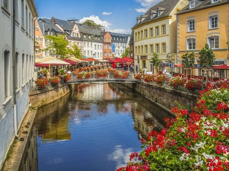 Steinbrücke mit roten und weißen Blumen über einem Kanal in Saarburgs Altstadt, mit bunten historischen Gebäuden und roten Sonnenschirmen.