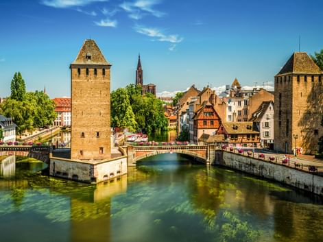 Zwei mittelalterliche Steintürme des Barrage Vauban im Wasser, verbunden durch eine Brücke in Straßburg. Historische Gebäude und Kirchturm sichtbar.