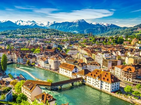 Panoramablick auf Luzerns Altstadt mit bunten Gebäuden entlang der türkisfarbenen Reuss, Holzbrücke und schneebedeckten Alpen im Hintergrund.