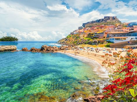 Sandstrand mit klarem türkisfarbenem Wasser in Castelsardo, Sardinien. Bunte Häuser erstrecken sich den Hügel hinauf, gekrönt von einer Festung.