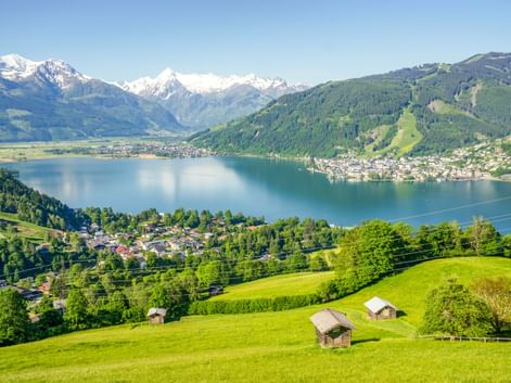 Panoramablick auf den Zeller See umgeben von grünen Hügeln und schneebedeckten Alpengipfeln. Traditionelle Holzhütten auf Wiesen im Vordergrund.