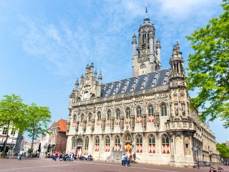 Gotisches Rathaus von Middelburg mit verzierter Fassade, hohem Turm und rot-weißen Fensterläden. Besucher auf dem Platz unter blauem Himmel.