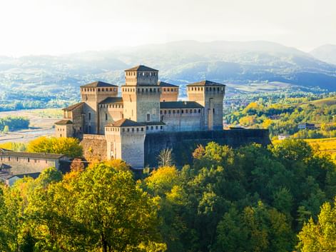 Burg Torrechiara auf einem bewaldeten Hügel, umgeben von grünen Bäumen mit sanften Hügeln und Bergen im Hintergrund bei weichem Licht.