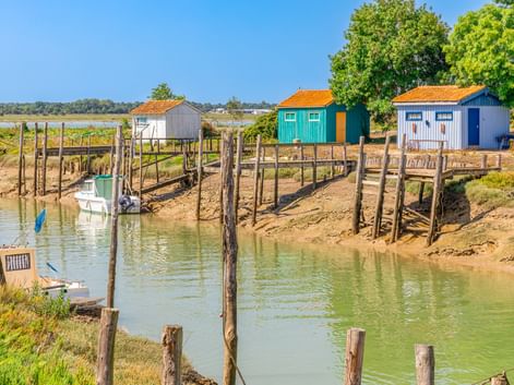 Kanal mit Holzpfählen und kleinem Boot, gesäumt von bunten Hütten mit orangen Dächern in Marennes, Frankreich, unter blauem Himmel.