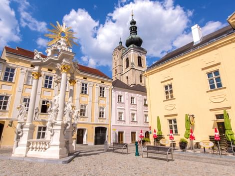 Barocker Platz in Krems-Stein mit verziertem weißen Monument, pastellfarbenen Gebäuden und Kirchturm unter blauem Himmel mit weißen Wolken.