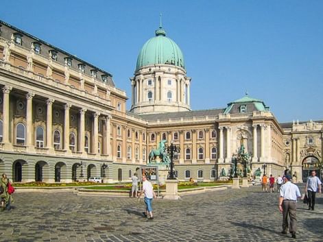 Burgpalast in Budapest mit seiner markanten grünen Kuppel und gelber Barockfassade. Besucher gehen über den Kopfsteinpflaster-Innenhof.