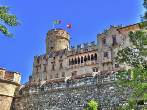 Schloss Buonconsiglio in Trient mit Steinmauern, zinnenbesetzten Türmen und italienischen Flaggen vor blauem Himmel, eingerahmt von grünen Bäumen.