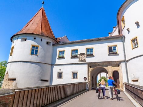 Zwei Radfahrer schieben ihre Räder über eine Brücke zum Eingang von Schloss Wörth. Weiße Schlossmauern mit rotem Turmdach unter blauem Himmel.