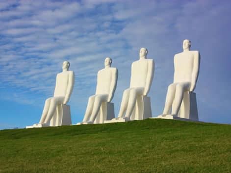 Vier monumentale weiße sitzende Figuren auf einem Grashügel vor blauem Himmel in Esbjerg. Die Skulpturen blicken zum Meer.