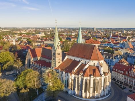 Luftaufnahme des Augsburger Doms mit zwei Türmen und roten Ziegeldächern, umgeben von historischen Gebäuden und Stadtlandschaft unter blauem Himmel.