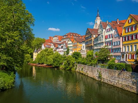 Bunte Fachwerkhäuser mit roten Dächern säumen den Neckar in Tübingens Altstadt. Ein Kirchturm ragt über den Gebäuden unter blauem Himmel empor.