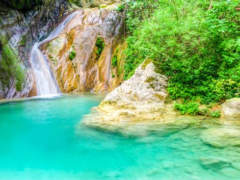 Wasserfall, der über goldbraune Felsen in ein türkisfarbenes Becken bei Nydri, Lefkada fließt. Üppige grüne Vegetation umgibt das natürliche Becken.