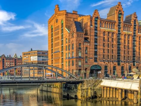 Rote Backsteingebäude der Hamburger Speicherstadt mit Bogenbrücke über Kanal. Historische Gebäude spiegeln sich im Wasser unter blauem Himmel.