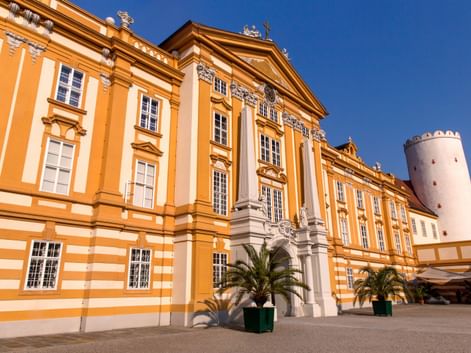 Barocke Fassade von Stift Melk in Gelb und Weiß mit prächtigem Eingangsportal. Palmen im Innenhof, weißer Turm rechts sichtbar.