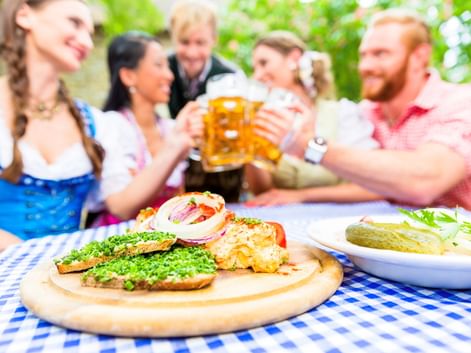 Bayerische Brotzeit auf einem Holzbrett mit Obatzda im Vordergrund. Menschen in Dirndl und Freizeitkleidung stoßen mit Bierkrügen an.
