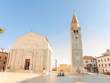 Sonniger Hafenplatz in Umag, Kroatien, mit weißer Kirche und steinernem Glockenturm. Palme links, Fußgänger auf dem Platz unter blauem Himmel.