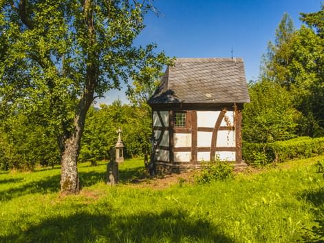 Fachwerkkapelle mit weißen Wänden und braunen Balken im Freilichtmuseum Roscheider Hof in Konz, umgeben von grüner Wiese und Bäumen unter blauem Himmel.
