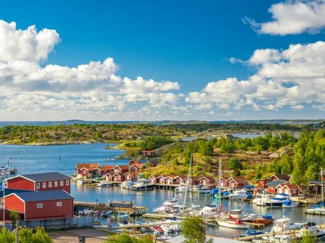 Hafen im Göteborger Schärengarten mit traditionellen roten Holzhäusern und Booten. Felsige Inseln mit grüner Vegetation unter blauem Himmel.
