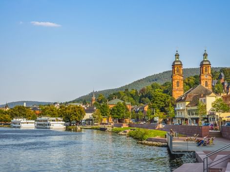 Blick auf Miltenberg mit den Zwillingstürmen der St. Jakobus Kirche am Main. Uferpromenade mit Booten, Hügel im Hintergrund.