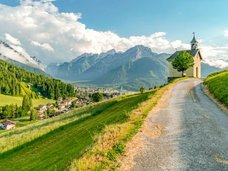 Kleine Kapelle mit Glockenturm an einem Schotterweg mit Blick auf Toblach und die Dolomiten unter blauem Himmel mit weißen Wolken.