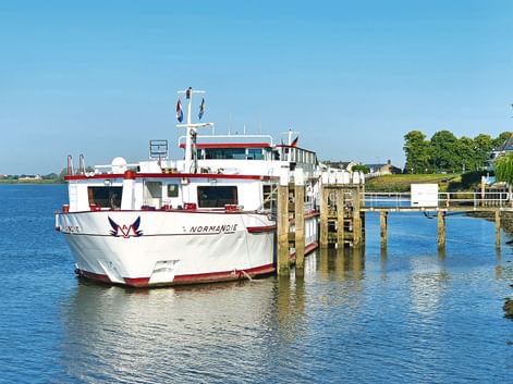 Weiß-rote Passagierfähre Normandie an einem Holzsteg in ruhigem blauen Wasser unter klarem Himmel, mit grünen Bäumen am Ufer.