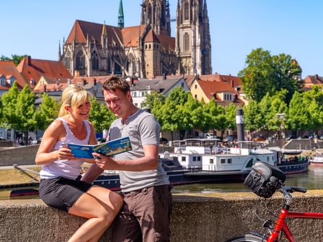 Zwei Radfahrer mit Karte am Donauufer, rotes Fahrrad daneben. Regensburger Dom mit Doppeltürmen erhebt sich über rotgedeckten Gebäuden.