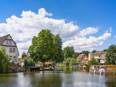 Wohnhäuser entlang eines ruhigen Wasserlaufs mit privaten Bootsstegen und Bootshäusern. Große Bäume rahmen die Szene unter blauem Himmel mit weißen Wolken.