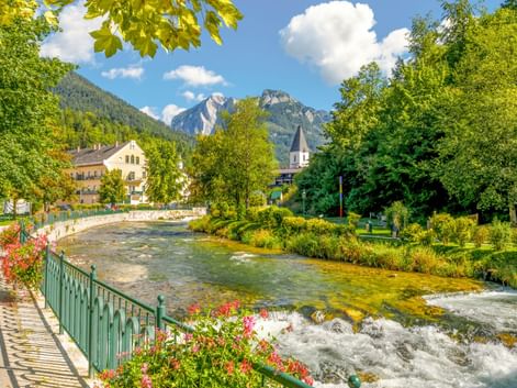 Fluss fließt durch Bad Aussee mit blumengeschmückter Promenade, Kirchturm und Bergen unter blauem Himmel mit weißen Wolken.