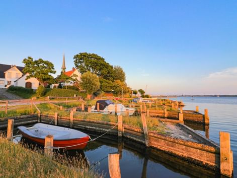 Holzsteg mit vertäuten Booten in Maasholm bei Sonnenuntergang. Kirche mit Turm und Häuser auf dem Hügel, ruhiges Wasser spiegelt den Himmel.