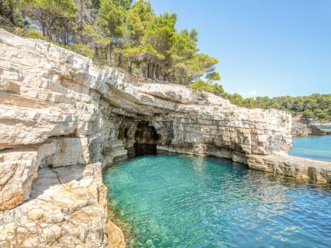 Natürliche Kalksteinhöhle mit türkisfarbenem Wasser in einer Bucht bei Pula. Weiße geschichtete Felsformationen mit grünen Bäumen unter blauem Himmel.