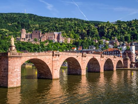 Historische Steinbrücke mit mehreren Bögen über den Neckar in Heidelberg. Ruinen des roten Sandsteinschlosses auf bewaldetem Hang sichtbar.