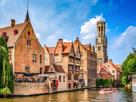 Mittelalterliche Backsteingebäude entlang eines Kanals in Brügge mit dem ikonischen Belfried im Hintergrund. Ein Touristenboot fährt auf dem Wasser.