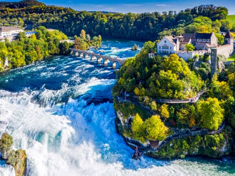 Luftaufnahme des Rheinfalls bei Schaffhausen mit tossendem Wasser, historischem Schloss, Steinbrücke und grünen Wäldern unter blauem Himmel.