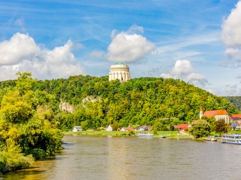 Die Befreiungshalle thront auf einem bewaldeten Hügel über Kelheim. Die Donau fließt unten mit Booten am grünen Flussufer.