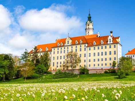 Benediktinerabtei in Donauwörth mit gelber Fassade und orangefarbenem Dach, umgeben von grüner Wiese mit gelben Löwenzahn und weißen Blumen.