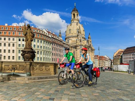 Zwei Radfahrer mit Tourenrädern und Packtaschen auf einem Kopfsteinpflasterplatz in Dresden, dahinter die Frauenkirche und historische Gebäude.