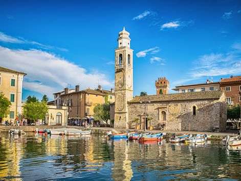 Kleiner Hafen in Lazise mit bunten Booten, historischem Glockenturm, Steingebäuden und mittelalterlichem Turm unter blauem Himmel.