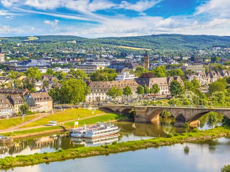 Luftaufnahme von Trier mit der Kaiser-Wilhelm-Brücke über die Mosel. Ein Passagierschiff liegt nahe der Brücke, dahinter die Stadt und Hügel.