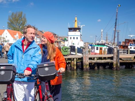 Zwei Radfahrer mit Lenkertaschen am Flensburger Hafen. Dahinter historische Boote, ein Leuchtturm und bunte Häuser unter blauem Himmel.
