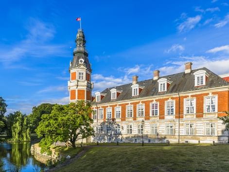 Schloss Hvedholm bei Faaborg mit orangefarbener Backsteinfassade, weißem Stuck und Uhrturm mit dänischer Flagge, umgeben von Rasen und Wassergraben.