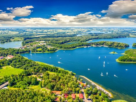 Luftaufnahme von Lötzen mit blauen Seen mit Segelbooten, grünen bewaldeten Halbinseln und vereinzelten Gebäuden unter blauem Himmel mit weißen Wolken.