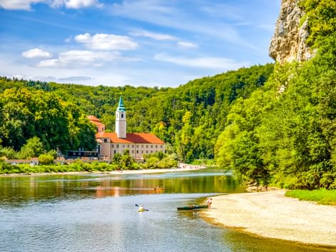 Kloster Weltenburg mit Glockenturm an der Donau, umgeben von grünen bewaldeten Hügeln. Kajakfahrer paddeln nahe einem Sandstrand im Vordergrund.