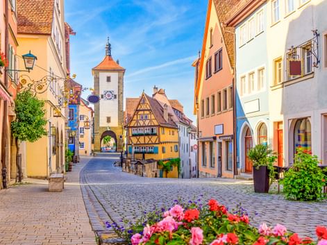 Kopfsteinpflasterstraße in Rothenburg mit bunten Fachwerkhäusern und Siebersturm mit orangefarbenem Dach. Rosa und rote Blumen blühen im Vordergrund.